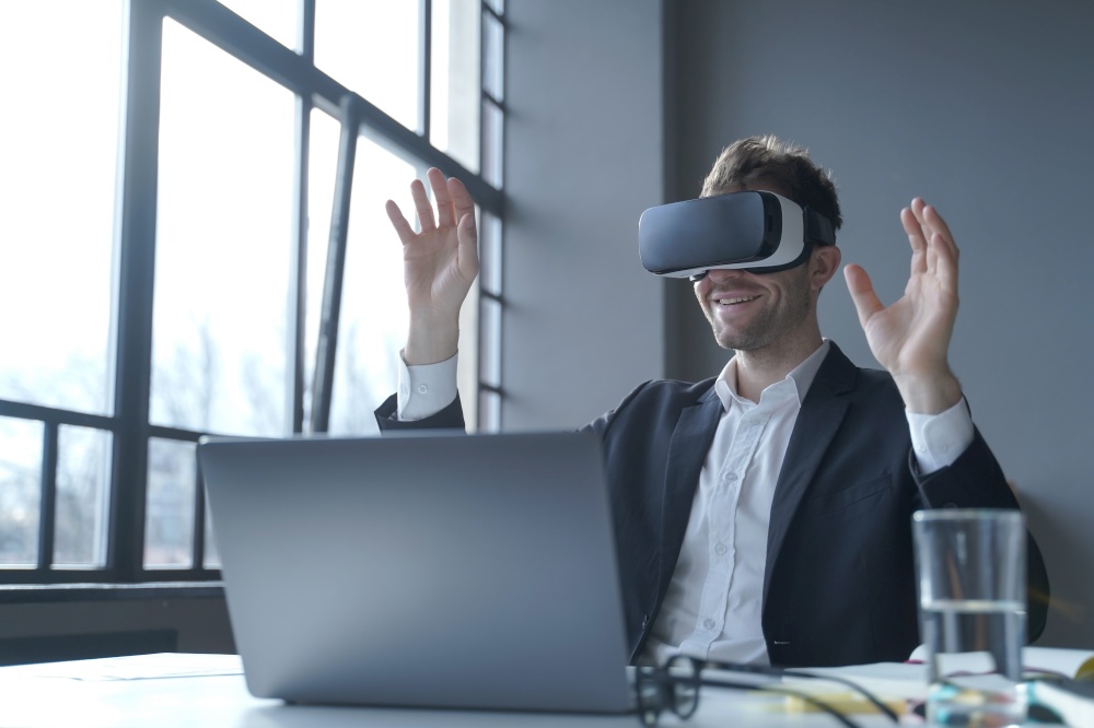 Employee experience with virtual reality. Young businessman in suit sitting at office table and using vr glasses goggles at work, male entrepreneur testing innovative method for business. Young businessman in suit sitting at office table and using vr glasses goggles at work