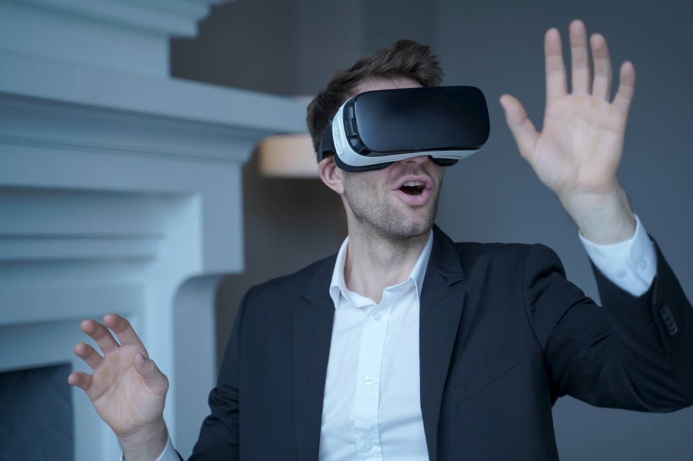 Man entrepreneur in formal suit sitting at home office while testing VR goggles, childishly impressed businessman using 3D technology first time with hands raised up trying to touch virtual objects. Man entrepreneur in formal suit sitting at home office while testing VR goggles