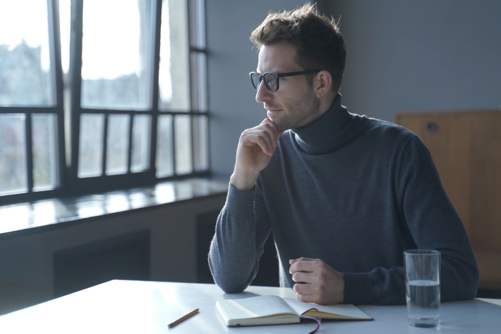 Young pensive thoughtful German man sitting at desk and looking out window while writing to do list or planing his working day. Handsome male writer in grey classic turtleneck pondering ideas. Young pensive thoughtful German man sitting at desk and looking out window