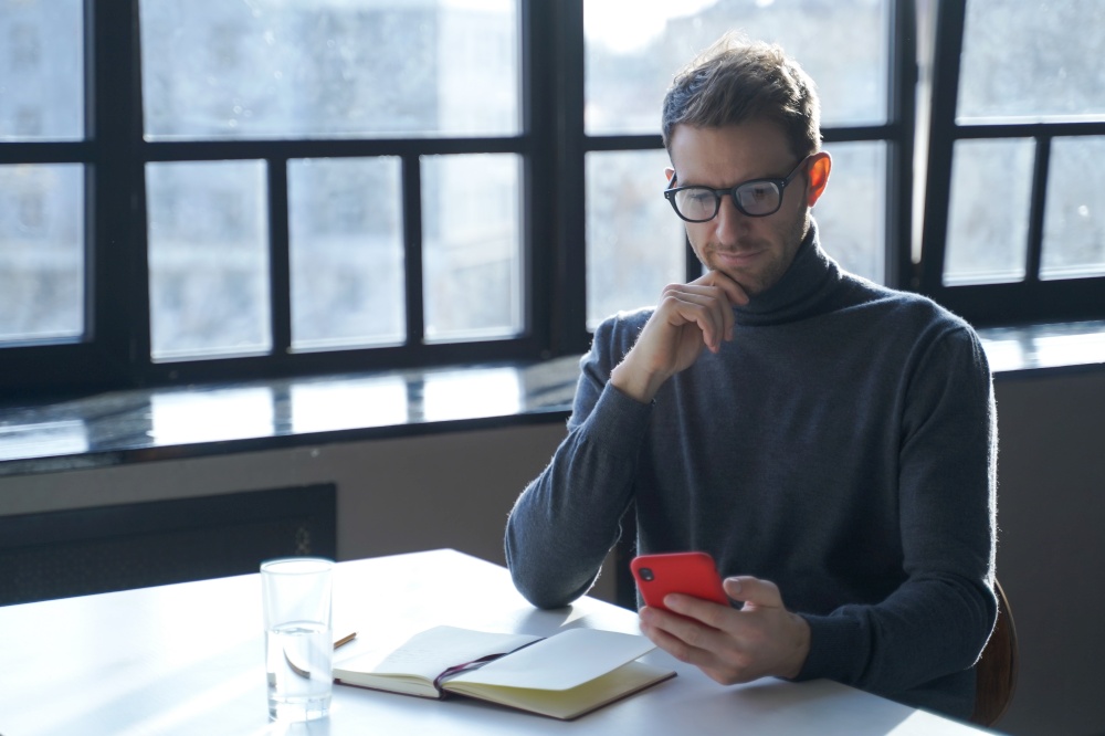 Young focused german businessman wearing glasses using mobile phone while sitting at desk with opened notebook, handsome austrian man holding smartphone, reading internet news or checking email. Young german businessman reading internet news or checking email on smartphone while sitting at desk