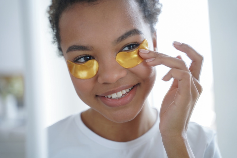 Happy afro girl is applying eye patches. Skin moisturizer with collagen serum. Beauty routine of lovely teenage african american girl. Mirror reflection of young spanish woman face. Morning skincare.. Happy afro teenage girl is applying eye patches. Skin moisturizer with collagen serum.