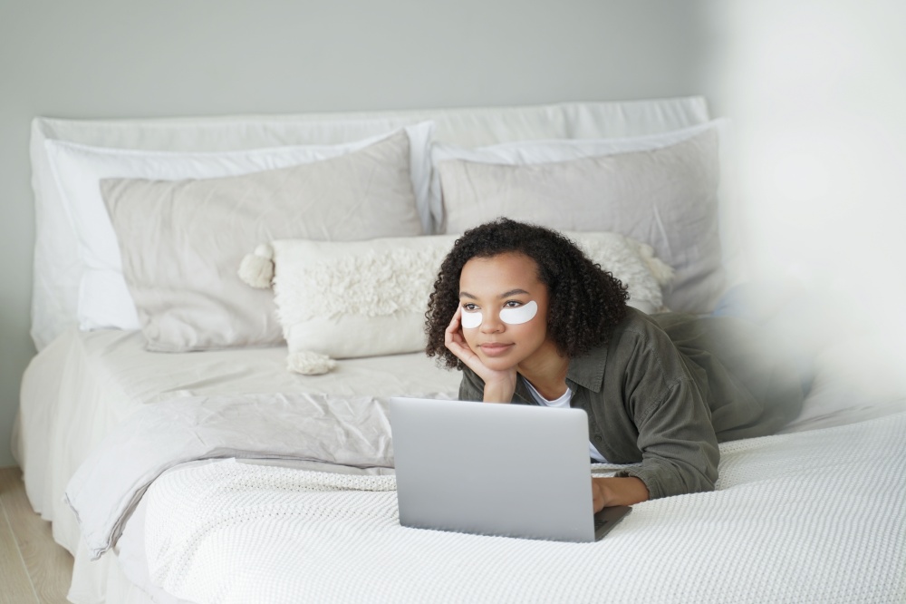 Weekend in bed. Lovely afro girl applies eye patches and relaxing at home. Girl is watching movie on laptop. Daily beauty routine of teenage girl. Cosmetic applying, hygiene and dermatology.. Weekend in bed. Lovely afro girl applies eye patches and relaxing at home watching movie.