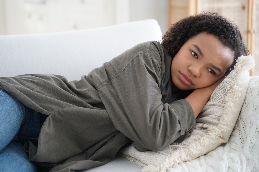 Frustrated lonely teen girl is lying on couch at home. Depressed african american teenager is suffering bullying or violence. Hispanic girl is desperate. Anxiety, stress and health problem concept.. Frustrated lonely teen girl is lying on couch at home. Teenager is suffering bullying or violence.