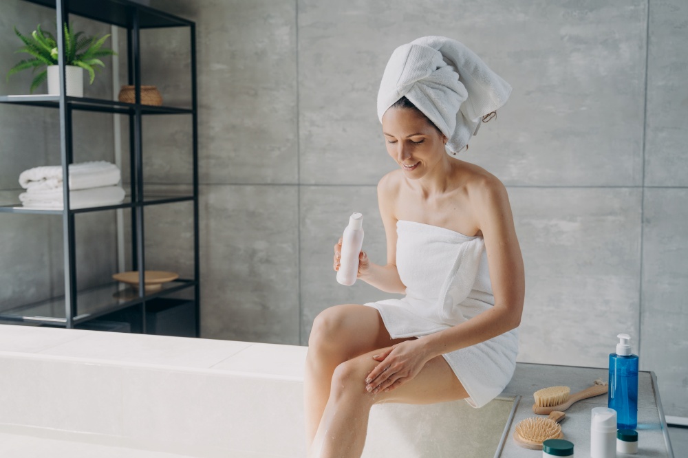 Attractive european woman wrapped in towel applying body lotion to her leg after shower. Young woman is bathing in morning at home. Anti-cellulite massage and bodycare. Enjoyment and relaxation.. Young woman is bathing in morning at home and applying body lotion to her leg after shower.