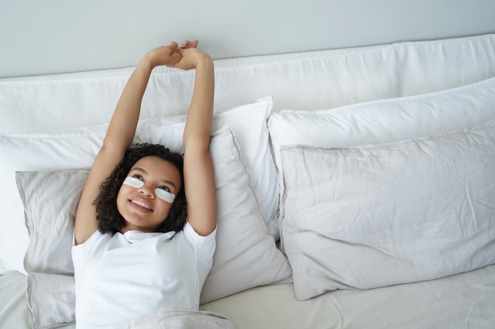 Young woman is stretching and relaxing in bed. Happy afro girl applies eye patches and relaxing at home enjoying weekend morning. Carefree spanish girl in bedroom. Comfort and leisure concept.. Happy young woman is stretching and relaxing in bed in morning. Comfort and leisure concept.