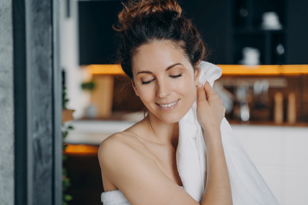 Young lady relaxing in bathroom, enjoying the bath and beauty routine. European girl is wiping face with towel after washing. Woman takes shower at home and doing skin care. Daily hygiene.. Young lady relaxing in bathroom, enjoying the bath and beauty routine. Girl wiping face with towel.