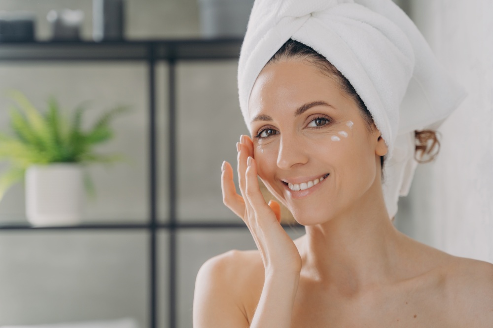 Face of beautiful european lady which applies cream. Attractive feminine caucasian girl wrapped in towel after bathing and hair washing. Happy young woman takes shower at home. Beauty routine.. Face of beautiful european lady which applies cream. Attractive feminine girl beauty routine.