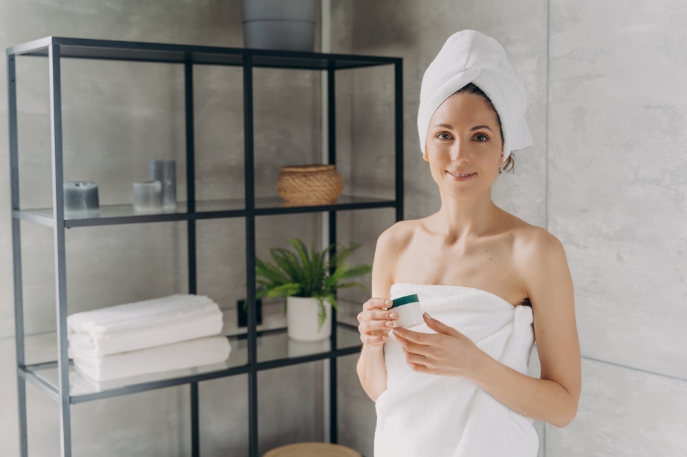 Attractive caucasian girl wrapped in towel after bathing and hair washing. European lady holding a cream jar. Happy young woman takes shower at home. Cosmetic applying, face and body care.. Attractive caucasian girl wrapped in towel after bathing and hair washing is holding a cream jar.