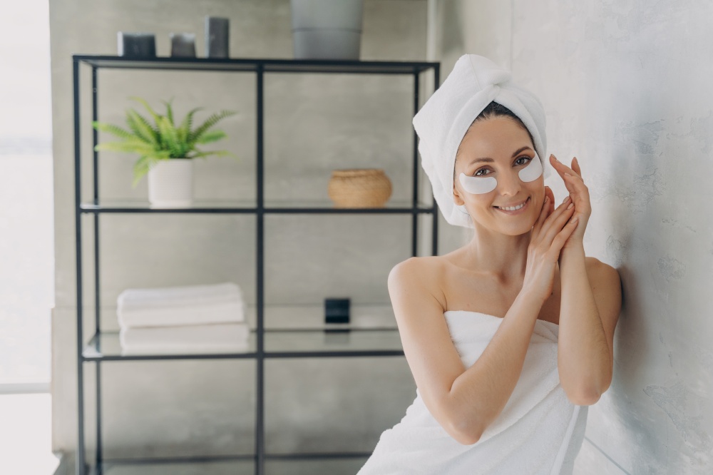 Young happy woman is relaxing at luxurious spa resort. Girl applies anti wrinkle eye patches. Morning beauty routine at home. Attractive european woman wrapped in towel after bathing.. Young happy woman is relaxing at luxurious spa resort. Girl applies anti wrinkle eye patches.