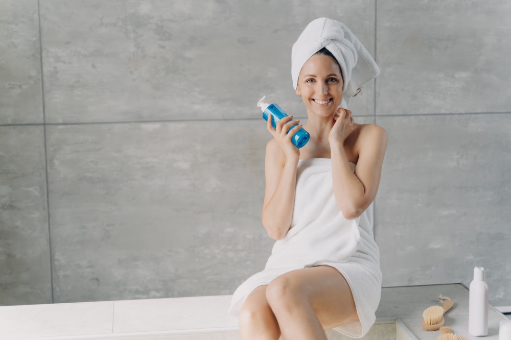 Face toner or micellar water advertising concept. Clean skin of beautiful caucasian woman wrapped in towel after bathing. Young happy woman takes shower at home holding face washing foam in bottle.. Young happy woman takes shower at home. Girl is holding face washing foam in bottle.