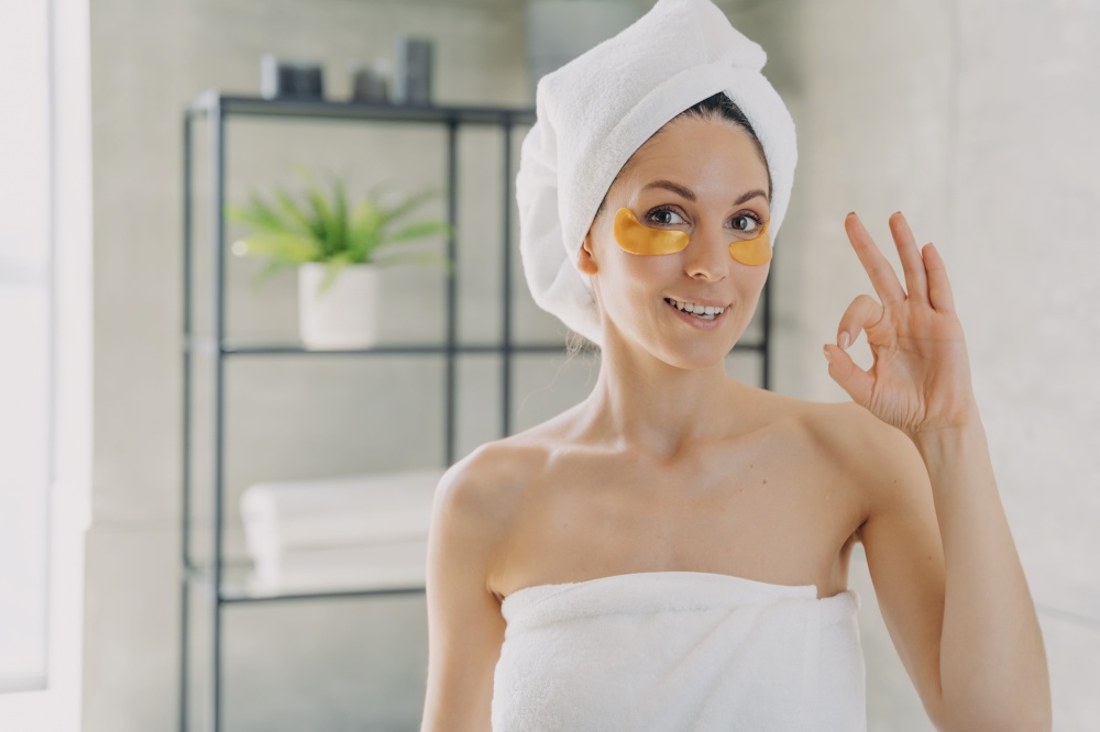 Girl applies collagen eye patches and gestures ok. Attractive woman wrapped in towel after spa procedures. Young hispanic lady does hair and body care. Relaxation at modern spa resort.. Girl applies collagen eye patches and gestures ok. Woman wrapped in towel after spa procedures