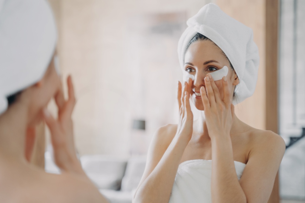 Girl applies eye patches and looking into mirror. Attractive european woman wrapped in towel after bathing. Young hispanic lady takes shower at home. Relaxation at spa resort. Beauty routine at home.. Girl applies eye patches and looking into mirror. Attractive woman wrapped in towel after bathing.