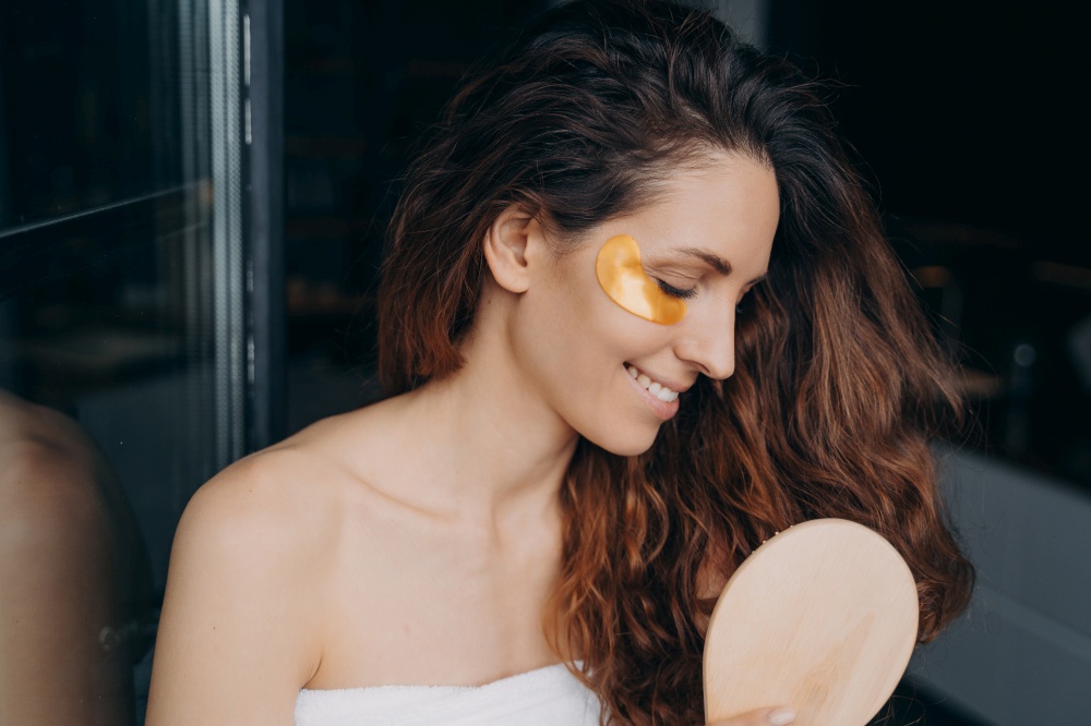 Young happy woman takes shower at home and doing daily hair and face care. Sweet brunette woman combs her soft curly hair with brush. Girl applying eye patches after bathing. Weekend morning at home.. Young happy woman doing daily hair and face care. Woman combs her hair and applies eye patches.