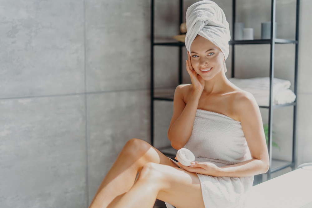Photo of young smiling European woman touches cheek, applies moisturizing cream on face, has happy expression, wrapped in soft bath towel, poses in bathroom. Beauty hygiene routine concept
