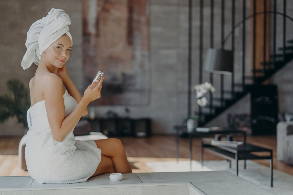 Indoor shot of good looking woman holds bottle of lotion has well cared smooth skin wears towel on head and around naked body after taking shower, poses against home interior. Beauty concept.