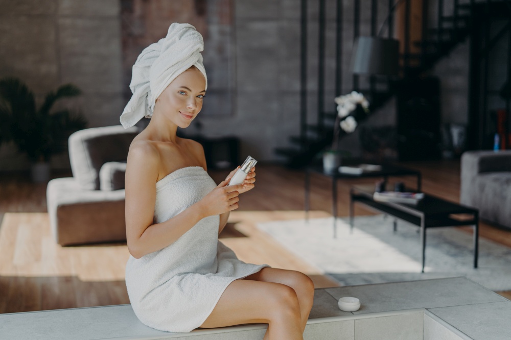 Horizontal shot of attractive lovely woman sits against cozy room background applies body lotion after taking bath uses cosmetic product for glowing skin wrapped in towel undergoes beauty treatments