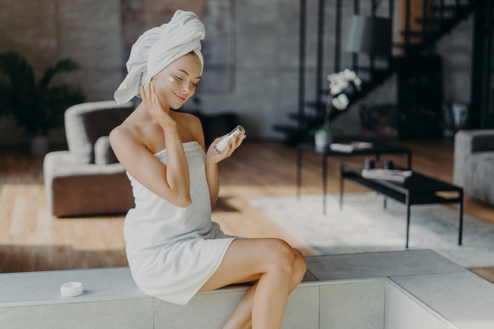 Smiling glad young woman touches healthy glowing skin, does daily beauty procedures at home, holds bottle of lotion, applies face cream, wrapped in bath towel, poses against cozy home interior