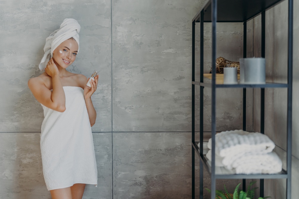 Pleased young European woman cares of her beautiful skin, applies moisturizer on face, holds bottle of lotion, stands against grey wall in bathroom, wrapped in bath towel, has refreshed look