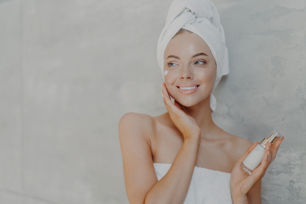 Pleased European woman puts on moisturizing cream on face, looks gladfully aside, touches face, has healthy glowing skin, poses against grey background with copy space for your advertisement