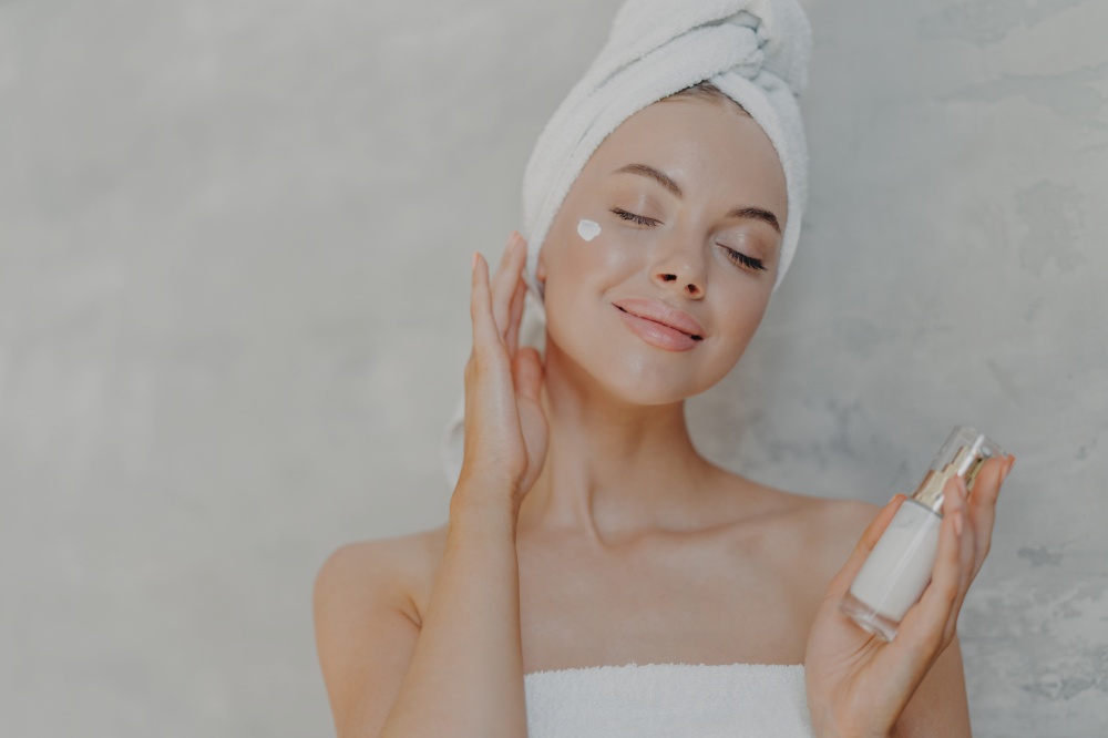 Headshot of pleased attractive woman applies face lotion, satisfied with new cosmetic product, keeps eyes closed, touches soft skin after bath, has well cared complexion, poses against grey wall