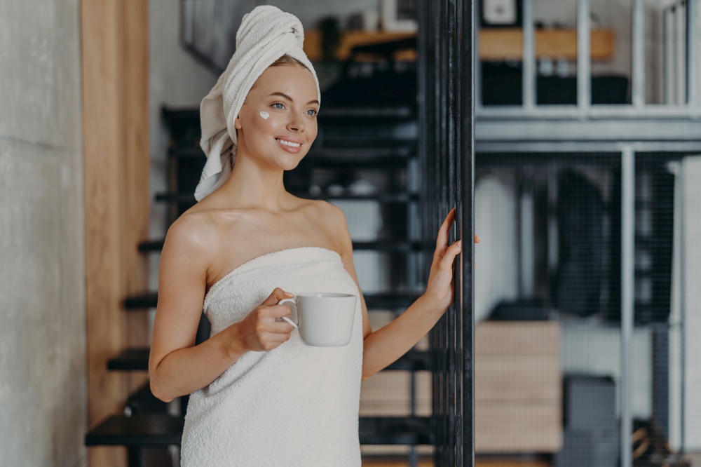 Photo of pleased smiling European woman enjoys day off at home, applies face cream, drinks tea, wrapped in towel, poses near strairs, thinks about something pleasant, has healthy glowing skin