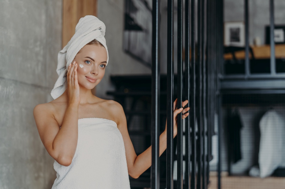 Beautiful healthy young woman applies face cream on complexion, looks thoughtfully somewhere into distance, wrapped in towels, stands indoor, has perfect pure skin. Women and cosmetology concept