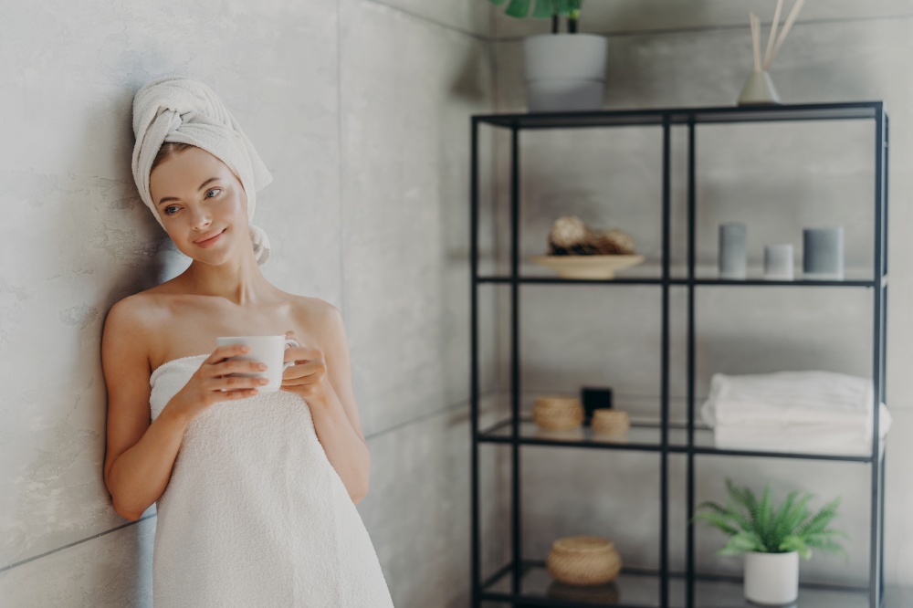 Photo of calm relaxed woman poses in towel against bathroom interior, wrapped in bath towel, drinks tea or coffee, has clean perfect skin, leans at grey wall. Beauty day, spa and skin care treatment