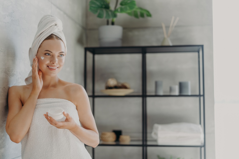 Lovely smiling Caucasian woman applies beauty cream on cheek, enjoys morning domestic skin care routine, wrapped in bath towel, grooming herself after showering poses in bathroom. Hygiene concept