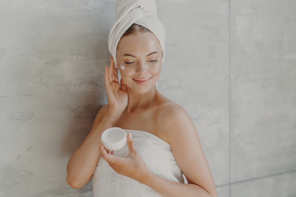 Grooming, cosmetology and wellness concept. Pleased female model poses with jar of moisturizing cream, applies cosmetic product on cheek, enjoys self care poses wrapped in towel near grey wall