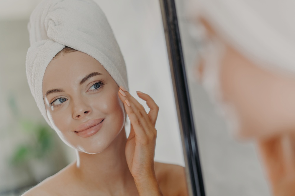 Beautiful woman touches face gently, looks at her reflection in mirror, enjoys softness of skin, stands with wrapped bath towel on head, has minimal makeup and manicure. Beauty treatment concept