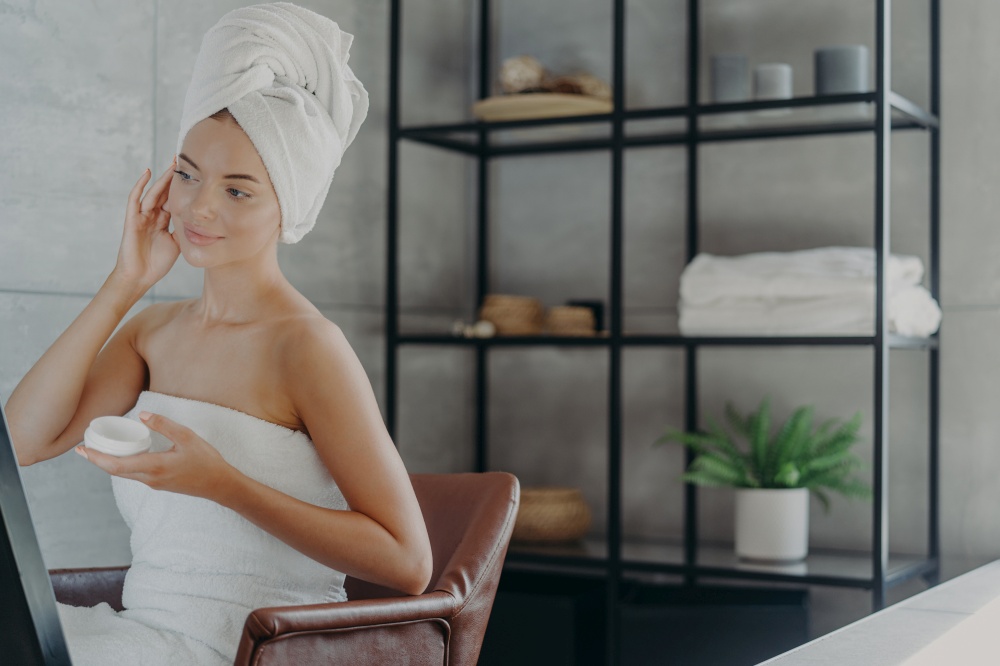 Attractive female model sits relaxed in armchair, applies cosmetic facial cream for healthy skin , looks into mirror, wrapped in bath towel, poses over bathroom interior, undergoes facial treatments.