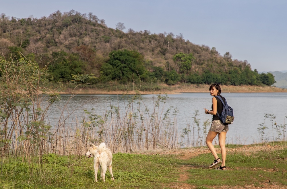 View of a young asia woman with white dog enjoying peaceful moment of beautiful view lake shore with mountains range in background. Pet and woman, Rest and enjoyment, lifestyle, Selective focus.