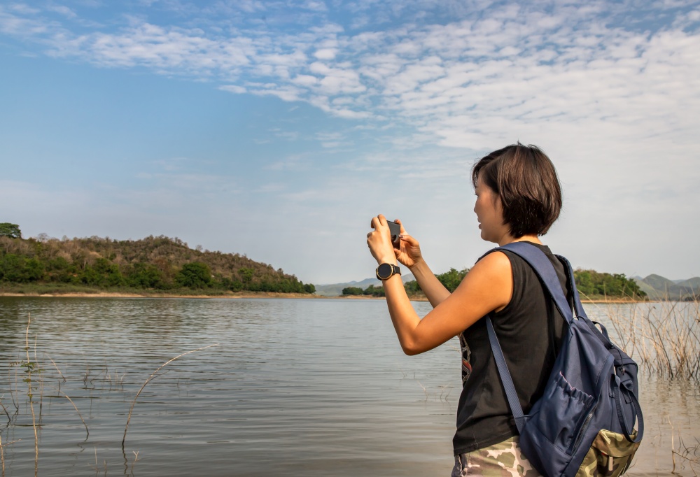 A side view of a young asia woman using her smartphone up and pointing it to take pictures view of lake shore with mountains range in background. Technology and holiday travel, lifestyle, Nature, No focus, specifically.