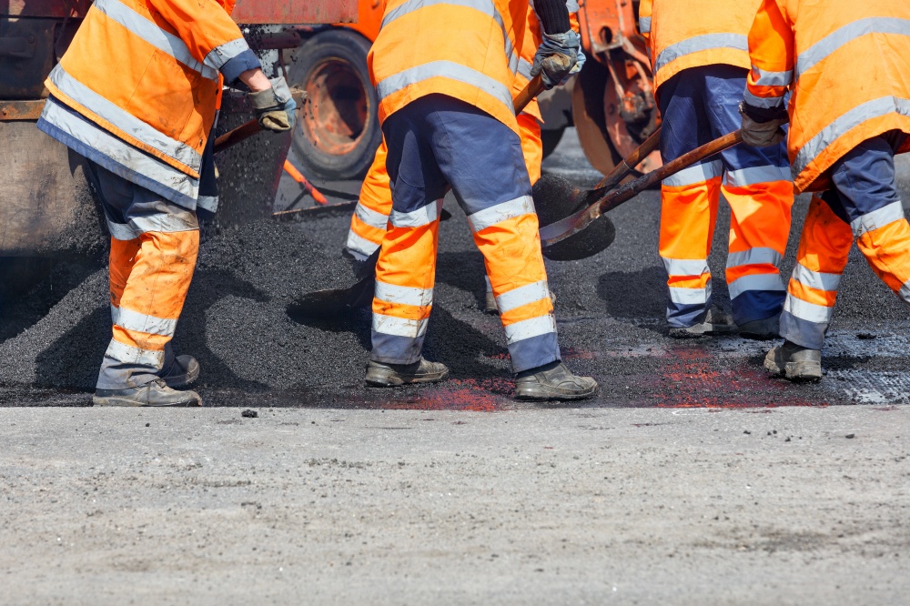 A team of road workers in orange uniforms resurfaces part of the road with fresh asphalt, spreading it with shovels, then compacting it with a road roller. Copy space.. A road worker is spreading fresh asphalt with shovels over the repair area to repair a section of road.