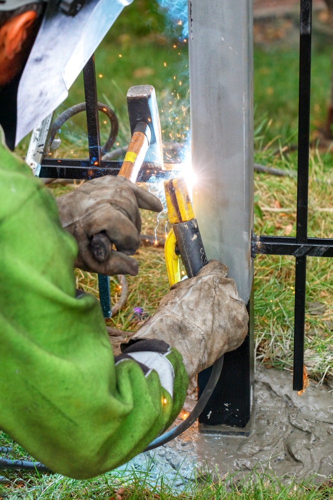 A welder in protective gloves welds a metal fence with an electrode, emitting bright sparks and blue smoke. Vertical image.. Bright sparks and flying blue smoke during electric welding of a metal fence by a worker in leather gloves.