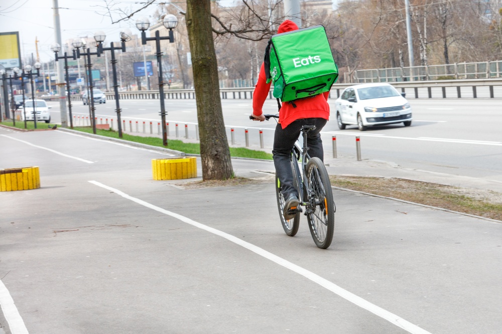 A courier with a green isothermal backpack behind his back rides along the sidewalk of a city street on a bicycle. Modern food delivery concept. Cpoy space.. Courier with an isothermal green backpack on his back on a food delivery bike on a city street.