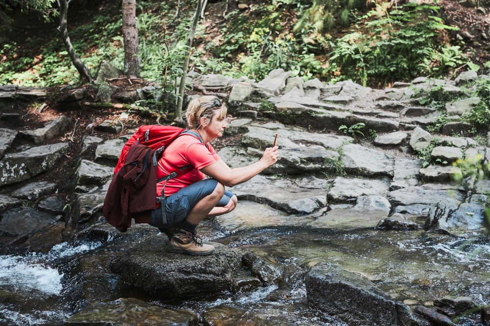 Taking pictures from vacation. Woman with backpack taking photos of landscape using smartphone camera standing on rock on mountain stream during summer trip
