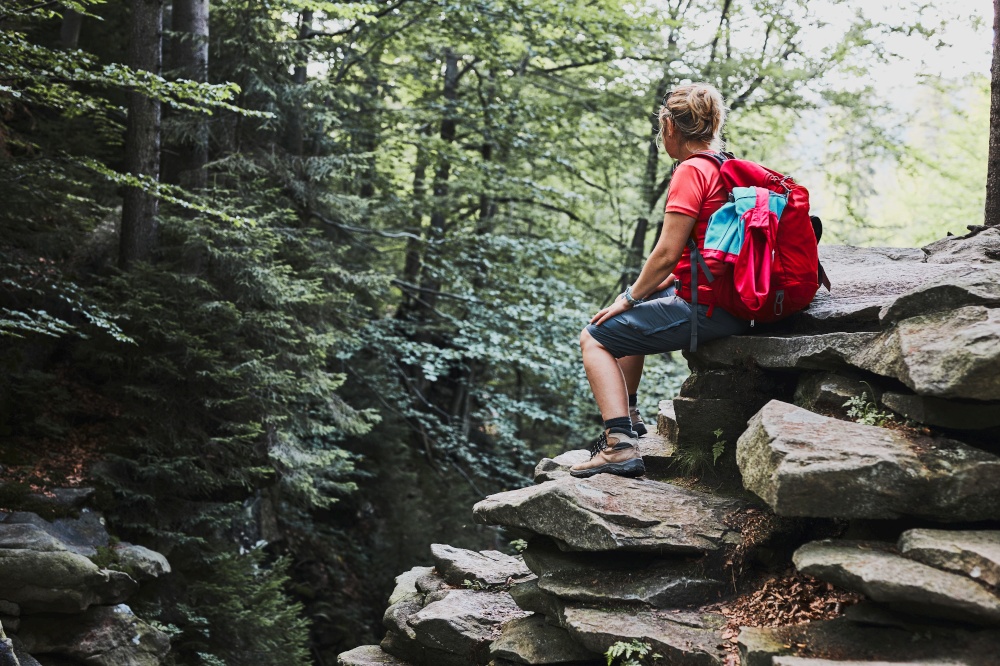 Woman with backpack hiking in mountains. Woman taking break sitting on rock. Spending summer vacation close to nature