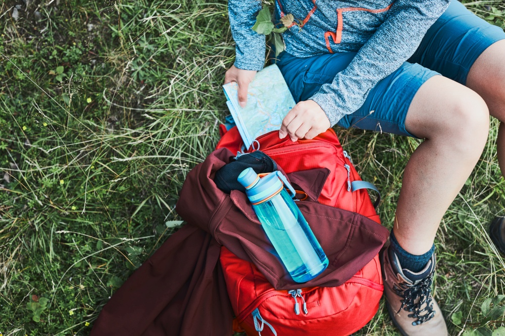 Woman with backpack having break during trip in mountains taking map out sitting on grass on summer vacation day