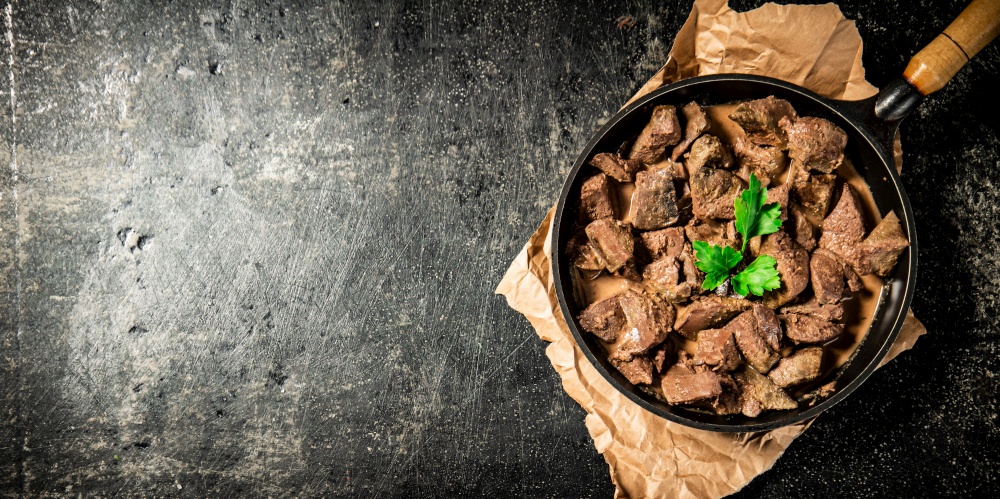 Liver in a frying pan on paper with a sprig of parsley. On a black background. High quality photo. Liver in a frying pan on paper with a sprig of parsley.