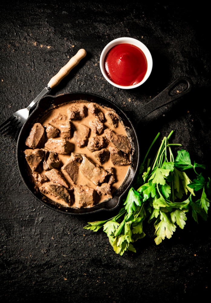Delicious fried liver in a frying pan with tomato sauce and parsley. On a black background. High quality photo. Delicious fried liver in a frying pan with tomato sauce and parsley.