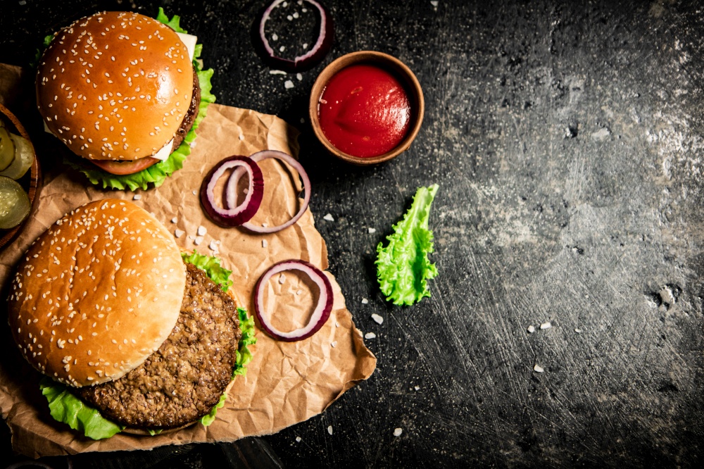 Burger on a cutting board with onion rings, tomato sauce and lettuce. On a black background. . Burger on a cutting board with onion rings, tomato sauce and lettuce.