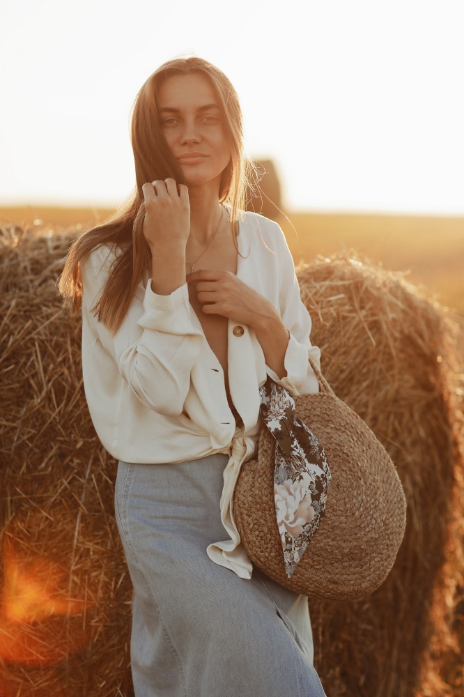 Full length portrait of a smiling beautiful brunette in a jeans skirt and straw bag in hand. Woman enjoying a walk in a wheat field with hay bales on summer sunny day. Full length portrait of a smiling beautiful brunette in a jeans skirt and straw bag in hand. Woman enjoying a walk in a wheat field with hay bales on summer sunny day.
