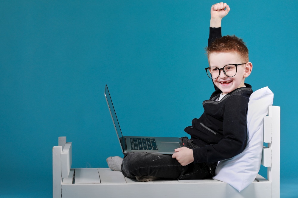 schoolboy in glasses using laptop sitting on bed. happy child doing school homework and joyfully raises his hands up. Online learning, distance lesson, education with online tutor at home isolation.. schoolboy in glasses using laptop sitting on bed. happy child doing school homework and joyfully raises his hands up. Online learning, distance lesson, education with online tutor at home isolation