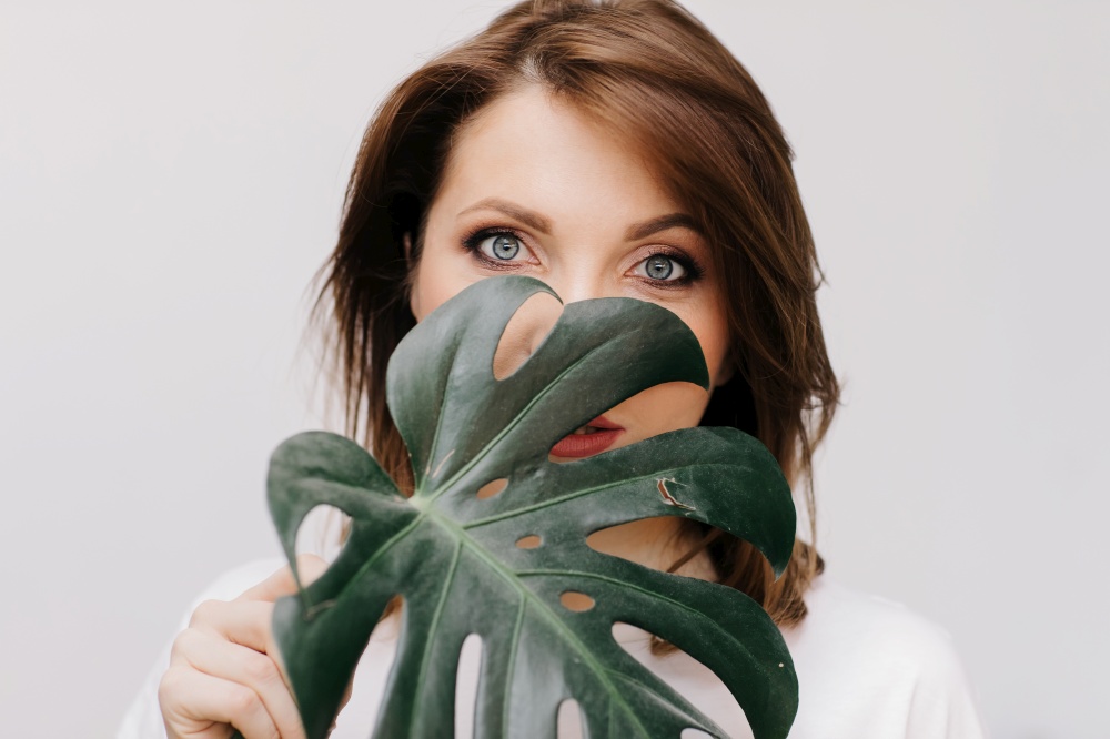 Portrait of young beautiful woman with makeup holds green tropical monstera leaf and covers part of her face. . High quality photo.. Portrait of young beautiful woman with makeup holds green tropical monstera leaf and covers part of her face. . High quality photo