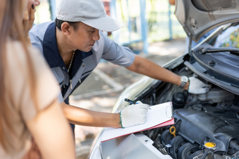 Asian male auto mechanic holds a clipboard and examines car engine breakdown problem with women customer and explain the root cause and estimate repair quotation, Car repair, and maintenance concept.