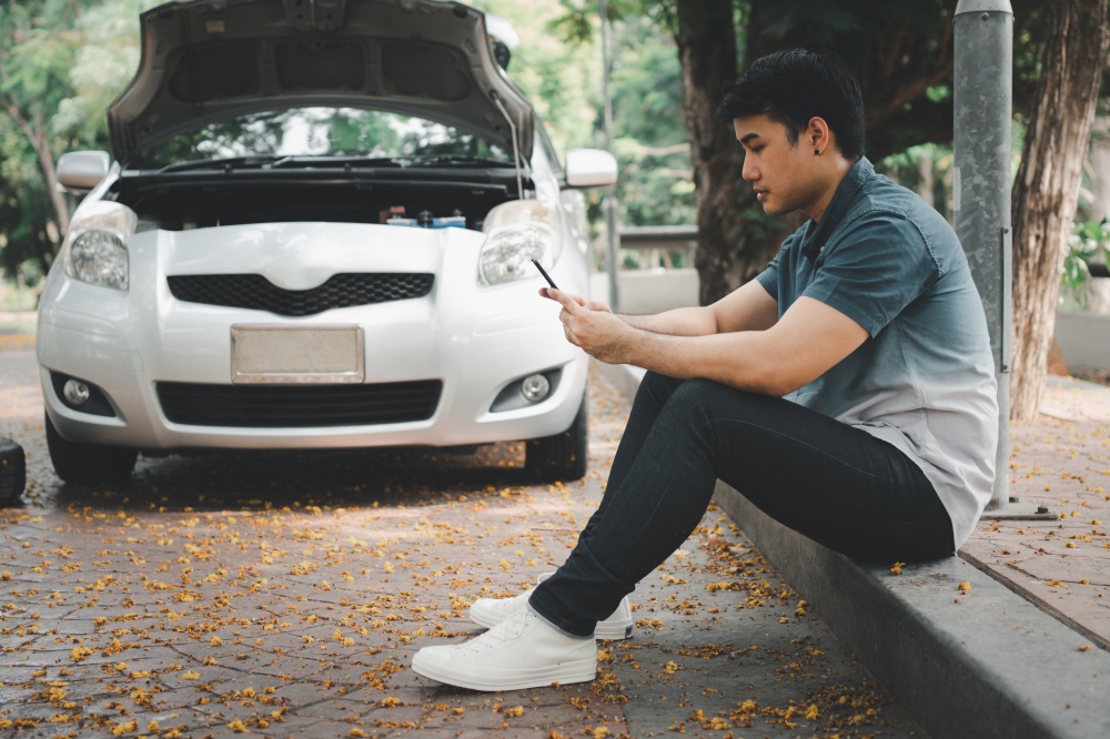 Asian man using smartphone for assistance after a car breakdown on street. Concept of vehicle engine problem or accident and emergency help from Professional mechanic