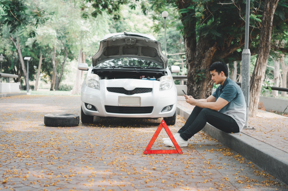 Asian man using smartphone for assistance after a car breakdown on street. Concept of vehicle engine problem or accident and emergency help from Professional mechanic