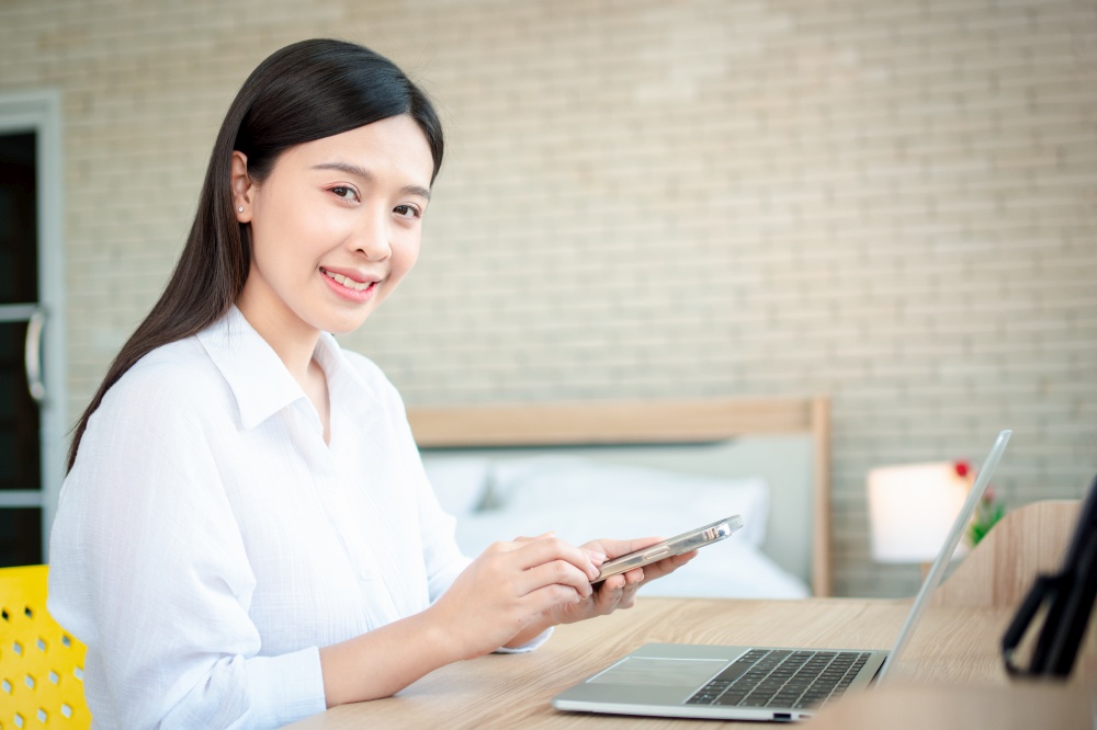 Happy beautiful asian woman working on a laptop and smartphone at the home office sitting at table. Happy female professional freelancer online using notebook pc and smartphone concept.