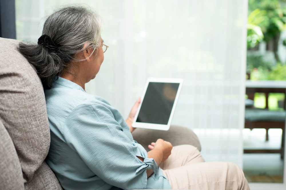 Senior asian woman wearing glasses and relaxing at home on a sofa and using tablet for reading news and e-book. Concept of technology for elderly.